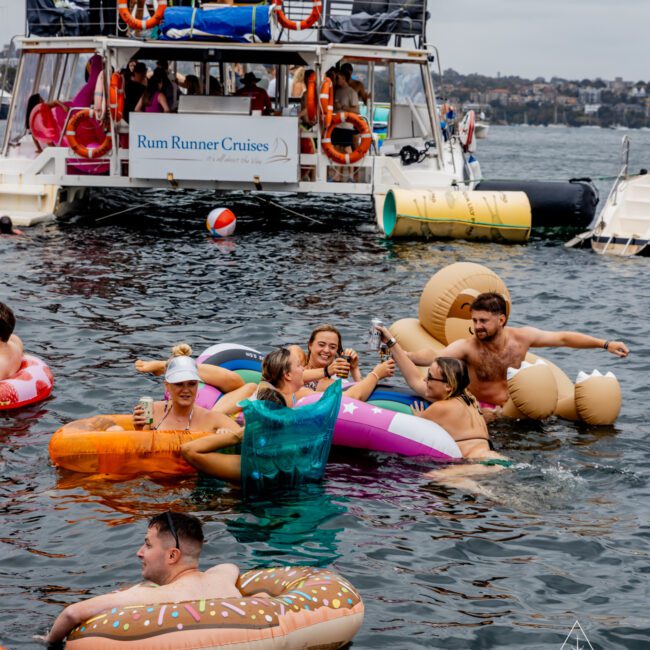 People in swimsuits are enjoying a party on a boat and in the water. Several are floating on inflatable pool toys, including a donut and a unicorn. The yacht in the background has a sign that reads "The Yacht Social Club Event Boat Charters" and is filled with more partygoers.