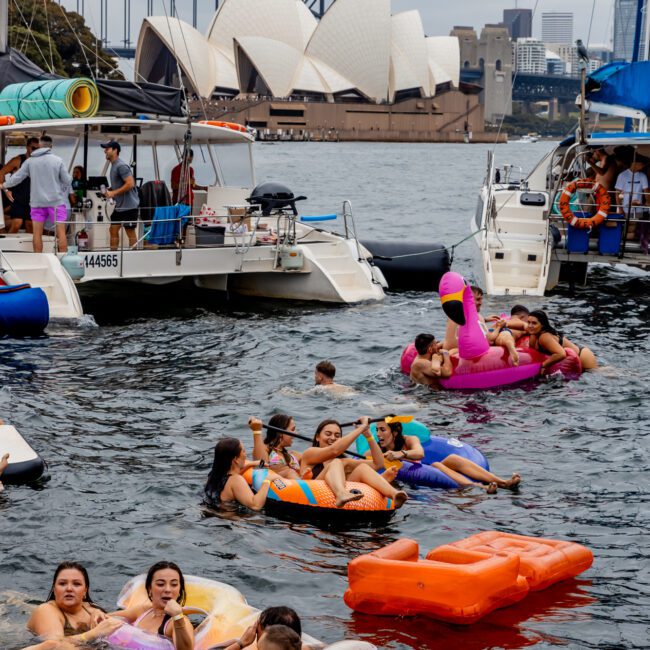 Experience a lively water party with people floating on colorful inflatables near luxury yachts. The Sydney Opera House and Harbour Bridge make a stunning backdrop as participants enjoy festivities under a cloudy sky, hosted by The Yacht Social Club Sydney Boat Hire.