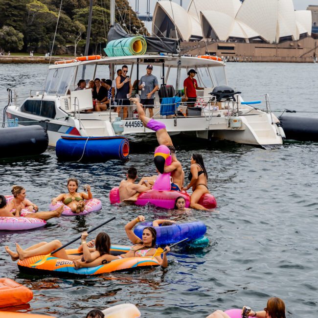 A group of people enjoy a sunny day on inflatable floats in the water near a docked boat. The iconic Sydney Opera House and Sydney Harbour Bridge are visible in the background. The scene is lively, with people relaxing and socializing at The Yacht Social Club event by Sydney Harbour Boat Hire.