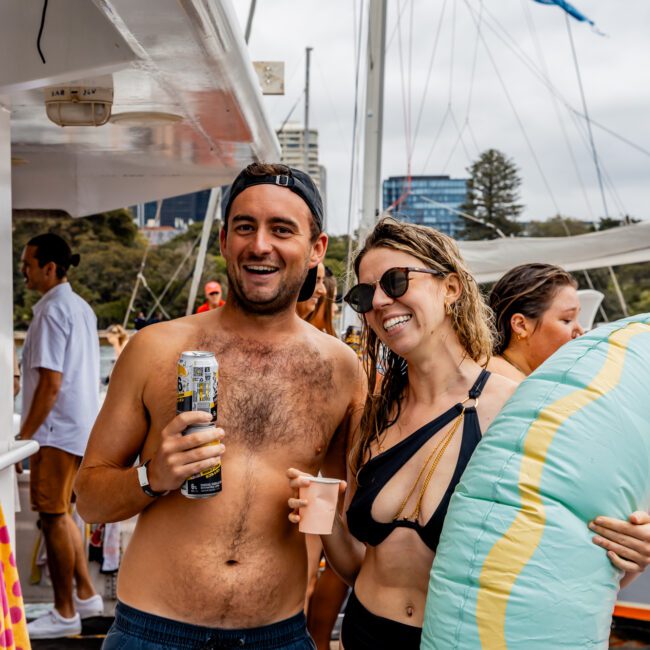 A shirtless man holding a beverage can and a woman in a bikini holding a floatie pose and smile on a boat crowded with people. The background shows more people socializing and a marina with yachts. It's a lively and sunny day at The Yacht Social Club Event, perfect for Boat Rentals and Parties in Sydney.