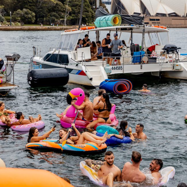 People enjoy a sunny day on a boat and in the water, floating on inflatables near a harbor. The Sydney Opera House and Sydney Harbour Bridge are visible in the background. Some are on colorful inflatable flamingos and other floaties, part of the lively Boat Parties Sydney The Yacht Social Club.