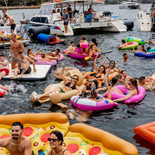 A fun scene of people enjoying the water on colorful inflatable floats, including pizza slices, a unicorn, and a flamingo. They are gathered near boats, with Luxury Yacht Rentals Sydney and The Yacht Social Club in view, while the iconic Sydney Opera House and Sydney Harbour Bridge provide a stunning backdrop.