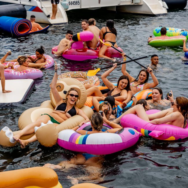 A lively group of people enjoying a summer day in the water with colorful inflatable rafts and pool floats, including a sloth, unicorn, and pizza slice. In the background, a boat from The Yacht Social Club floats with more people on board, indicating a fun and festive atmosphere.