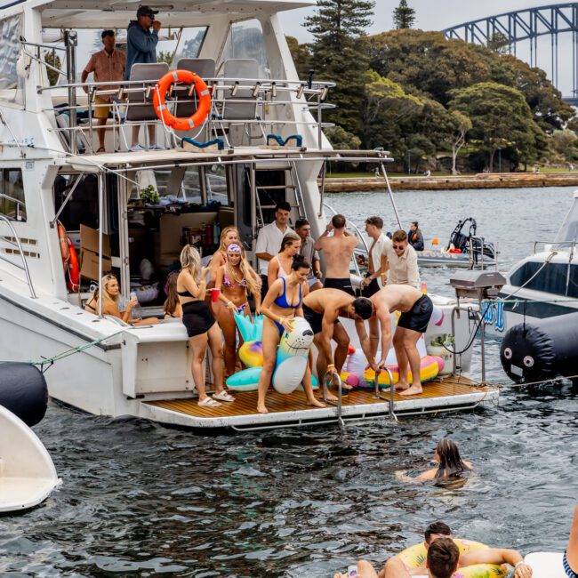 A group of people in swimwear enjoy a day on a luxury yacht docked on a waterway. Some are on the boat while others float nearby on inflatable toys. The scene is lively with a backdrop of trees and a bridge in the distance, capturing the essence of The Yacht Social Club Sydney Boat Hire experience.