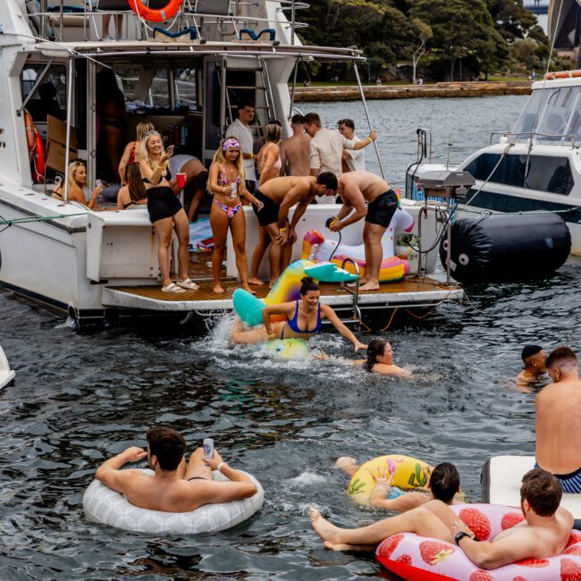 People are enjoying a sunny day on a boat docked by the shore. Some are lounging on floats in the water, while others are socializing on the boat from The Yacht Social Club Sydney Boat Hire. Colorful floaties with various designs are in use. A bridge and trees are visible in the background.