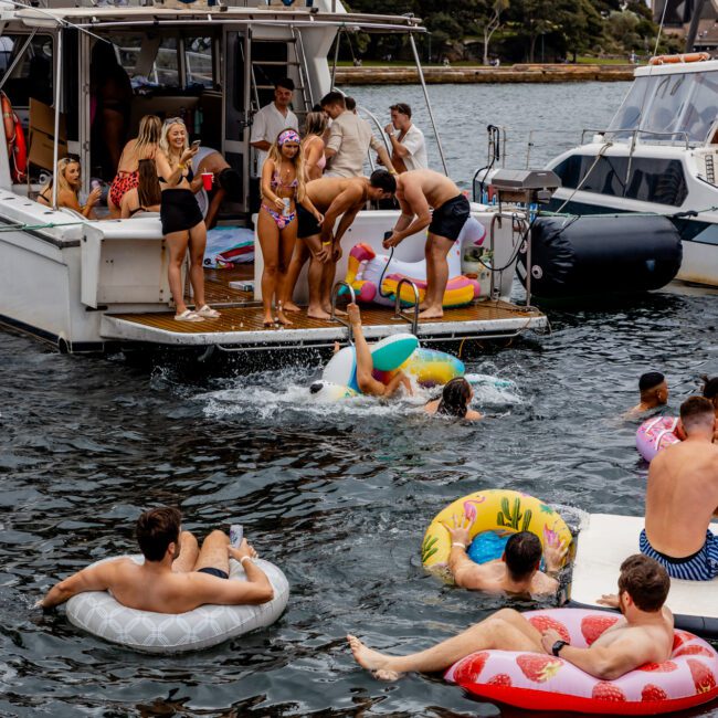 A group of people enjoying a party on the water near boats. Some are lounging on inflatable floats in the water, while others are on the deck, laughing and interacting. A bridge and trees are visible in the background under a cloudy sky—an ideal scene for Boat Parties Sydney The Yacht Social Club.