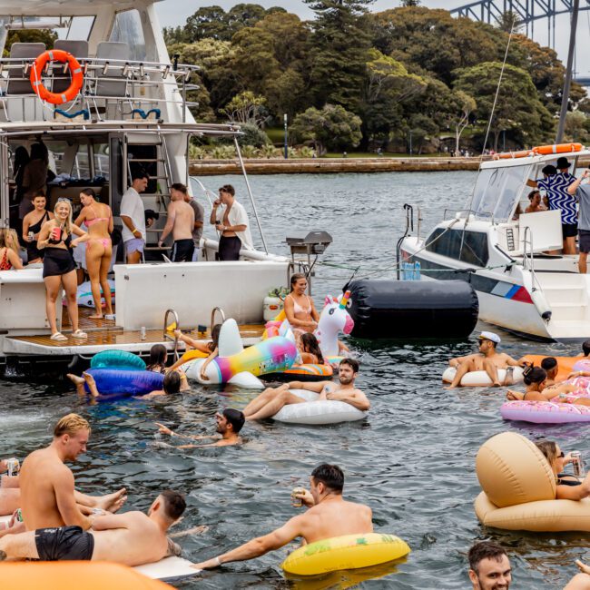 People are enjoying a party on a lake with various inflatables, including unicorns and tubes. Some are on boats from Sydney Harbour Boat Hire The Yacht Social Club, while others swim and float in the water. A bridge and lush greenery are visible in the background. The scene is lively and festive.