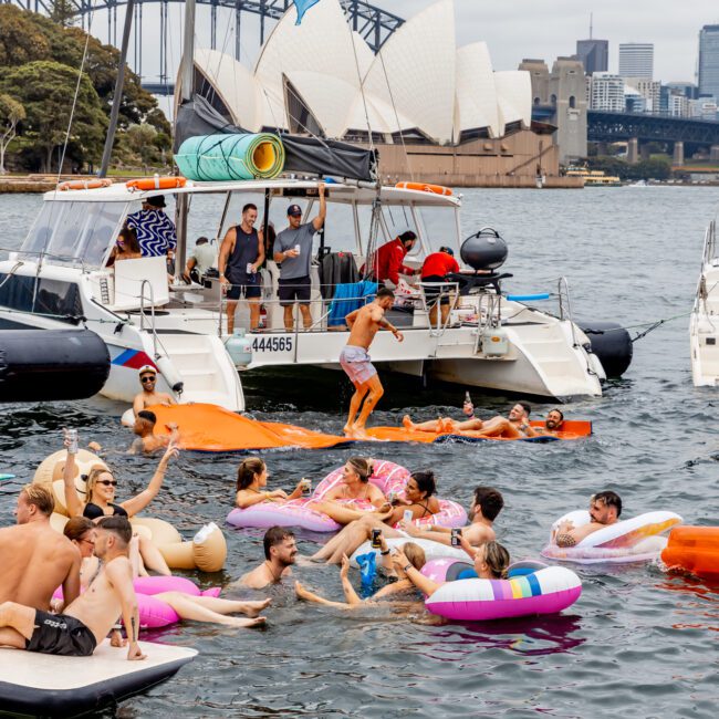 A group of people are enjoying a day on the water near the Sydney Opera House and Sydney Harbour Bridge. Some are floating on colorful inflatables, while others are on a catamaran. Despite the overcast sky, the atmosphere is lively and festive, reminiscent of Boat Parties Sydney The Yacht Social Club events.