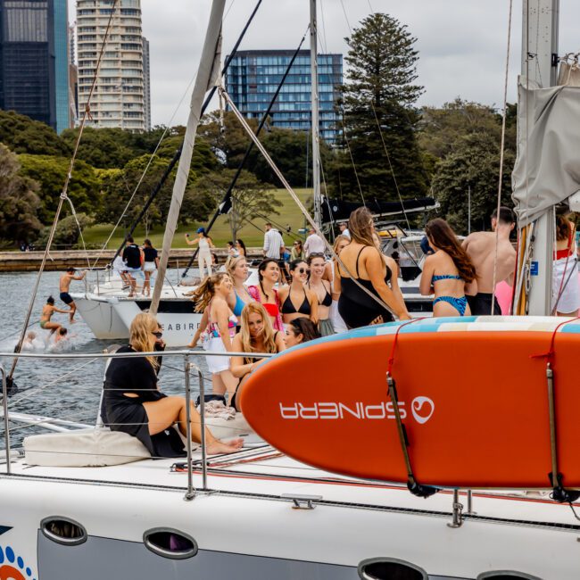 A group of people in swimsuits relaxes and socializes on the deck of a sailboat docked in a harbor. An orange paddleboard is secured on the side of the boat. This is The Yacht Social Club event by Luxury Yacht Rentals Sydney. Trees, modern buildings, and other boats are visible in the background under an overcast sky.