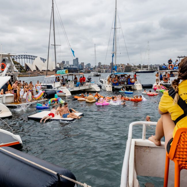 People are gathered on boats and in the water, enjoying a social event hosted by The Yacht Social Club Sydney Boat Hire. Some are lounging on inflatable floats. A lifeguard in a yellow shirt sits on a boat, overseeing the scene. The backdrop features a city skyline and an overcast sky.