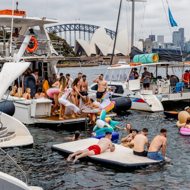 A lively scene on the water shows people enjoying The Yacht Social Club Event near the Sydney Opera House and Harbour Bridge. Partygoers are on boats and colorful inflatables in the water, socializing and having fun. The sky is overcast, but the atmosphere is festive.