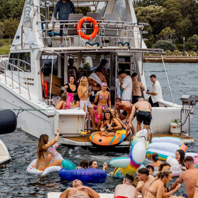 People are enjoying a lively boat party in a scenic area. The Yacht Social Club Event Boat Charters is packed with party-goers, and many people are in the water on inflatable floats. The atmosphere is festive and relaxed, with many participants wearing swimwear and smiling. Trees and greenery are in the background.