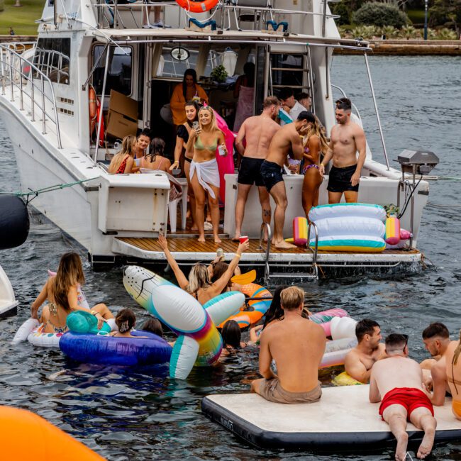 A lively scene of people enjoying a day on and around a yacht in a lake or river, reminiscent of Boat Parties Sydney The Yacht Social Club. Some are lounging on inflatable floats, while others are standing or swimming near the yacht. There's a mix of colorful pool floats and the environment is cheerful and relaxed.