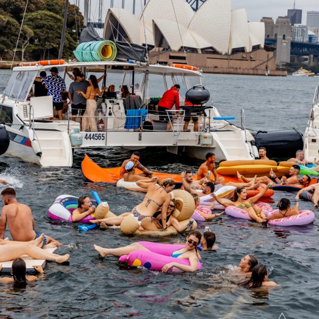 A lively group of people enjoy a boat party near the Sydney Opera House, hosted by The Yacht Social Club. Some are on the boat, while others float in various inflatable pool toys in the water. The atmosphere is fun and social, with music and laughter filling the scene, set against sailboats and the Sydney Harbour Bridge.