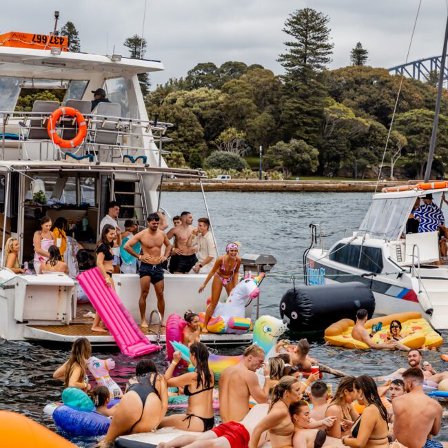 A lively scene of people enjoying a boat party hosted by The Yacht Social Club Sydney Boat Hire. Some are on the boat, while others float on colorful inflatables or lounge in the water. Everyone is in swimwear, and the background shows trees and part of a bridge. The atmosphere is festive and fun.