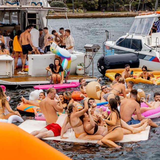 A lively group of people enjoy a summer day on a lake, with many floating on inflatables like unicorns and pink doughnuts. Several Luxury Yacht Rentals Sydney are docked nearby, and people are engaging in social activities, dancing, and relaxing in swimwear.