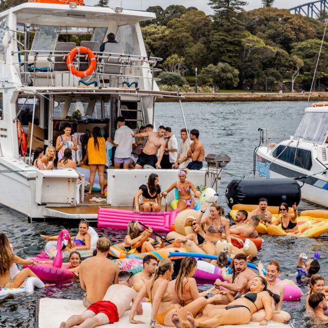 A lively scene of people enjoying a sunny day on the water with Luxury Yacht Rentals Sydney. Various individuals are gathered around and on a yacht, some swimming, and many lounging on inflatable floaties. The background features greenery and part of a bridge.