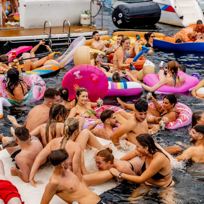 A vibrant pool party scene with people having fun on inflatables and in the water. Several individuals are gathered on a floating platform, while others enjoy colorful floaties like flamingos and donuts. In the background, luxury yachts from Boat Rental and Parties Sydney The Yacht Social Club are docked.