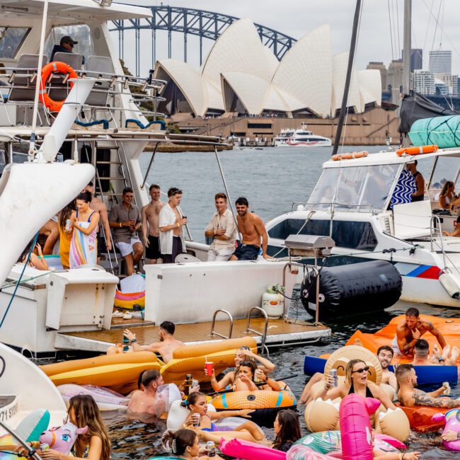 A lively boat party hosted by The Yacht Social Club on the water, with numerous people relaxing on colorful inflatables near boats. In the background are the Sydney Opera House and the Sydney Harbour Bridge. The scene is vibrant and festive, indicating a sunny gathering.