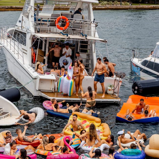 A lively group enjoys a party on and around a large boat docked in Sydney Harbour. Some float nearby in colorful inflatable rings and loungers, while others socialize on the boat. The scene is festive with a variety of floats, including a unicorn and flamingo, part of The Yacht Social Club Event Boat Charters.