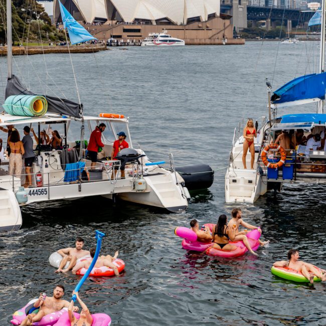 People enjoying a festive atmosphere on inflatable floaties and yachts in a harbor, with the Sydney Opera House and Sydney Harbour Bridge visible in the background. The water is filled with colorful inflatables, and some people are on the decks of luxury yacht rentals by The Yacht Social Club.