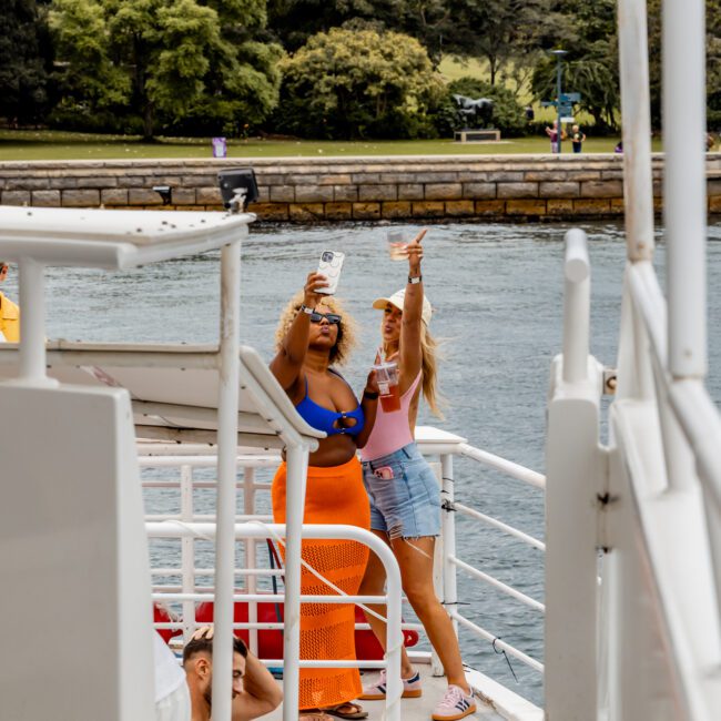 Two people on a boat from The Yacht Social Club are taking a selfie near the water. One is wearing a blue top and orange skirt, while the other is in a pink top and shorts. Other passengers on the boat are seen in the foreground. Trees and a walkway are visible in the background.