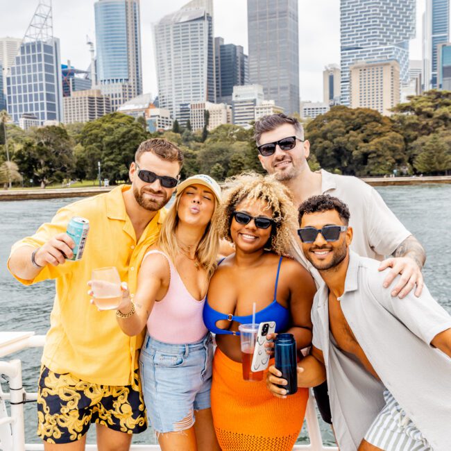 A group of five friends poses cheerfully on a boat with a city skyline in the background. They are dressed in casual summer attire, holding drinks, and smiling at the camera. Tall buildings and green trees are visible in the distance under a cloudy sky, courtesy of The Yacht Social Club Sydney Boat Hire.