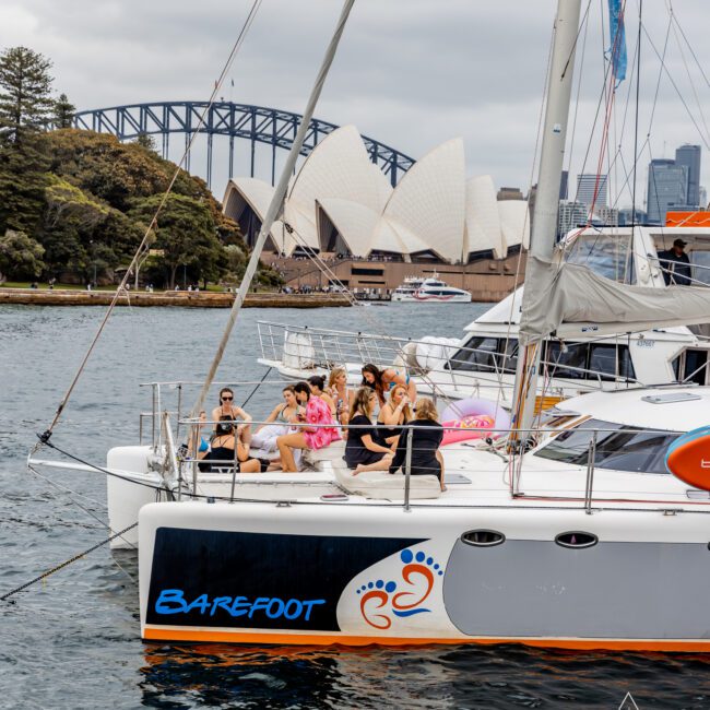 A group of people wearing festive outfits and sunglasses are gathered at the front of a catamaran named "Barefoot." They are sailing in a harbor with the Sydney Opera House and Sydney Harbour Bridge visible in the background under a cloudy sky, enjoying their time with The Yacht Social Club Sydney Boat Hire.