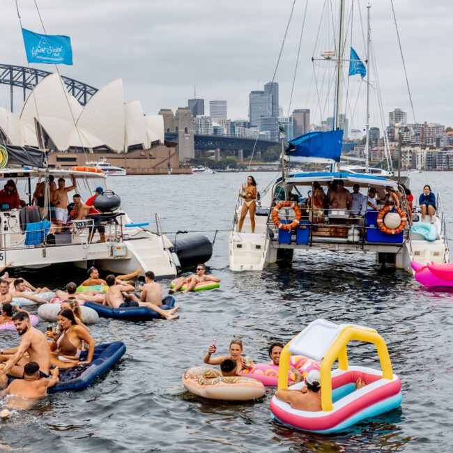 A lively scene of people lounging on inflatable floaties and swimming between two anchored boats. Passengers on board enjoy the day with flags flying. In the background are the iconic Sydney Harbour Bridge and Sydney Opera House. The Yacht Social Club Event Boat Charters adds a touch of elegance to the event.