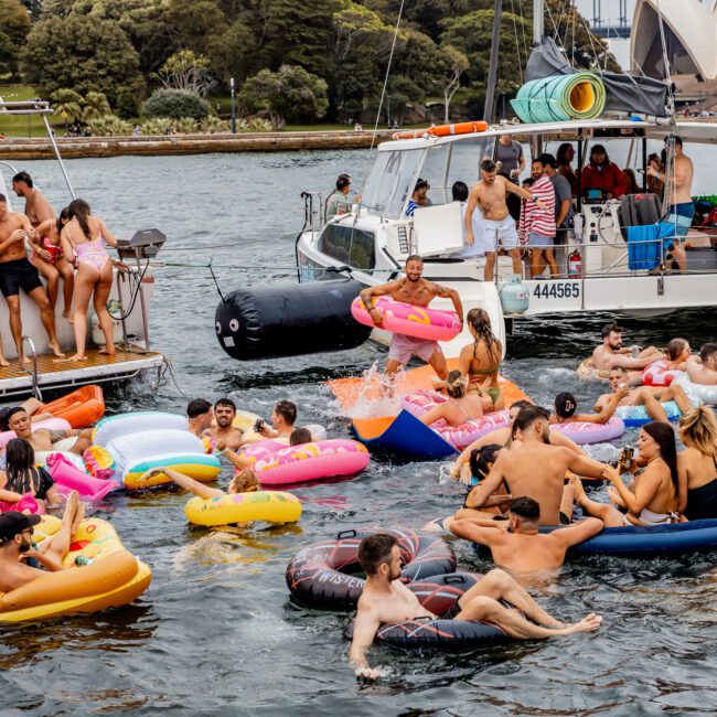 A lively group of people enjoys a day on the water with colorful floaties, while two boats from The Yacht Social Club Sydney Boat Hire are anchored nearby. The group, including adults and children, has fun under a gray, cloudy sky. A bridge and trees are visible in the background.