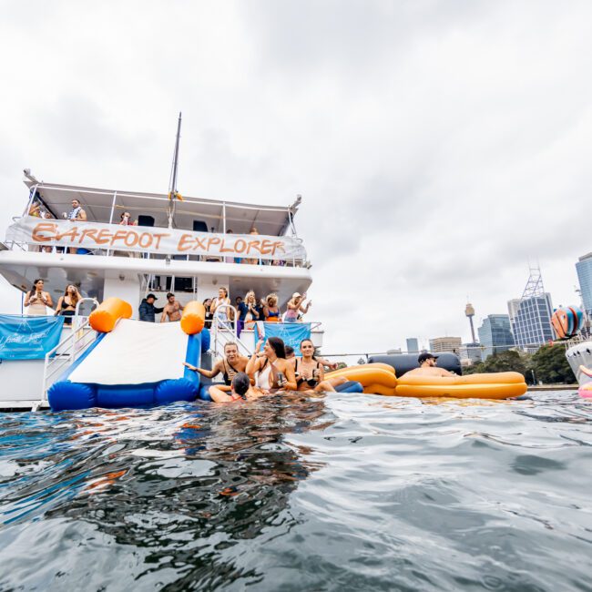 A group of people enjoy a sunny day at sea near a large yacht named "Barefoot Explorer," courtesy of The Yacht Social Club Event Boat Charters. Some are on the yacht's deck, while others float on inflatable pool toys and swim in the water. The sky is partly cloudy, and nearby boats are anchored in the background.