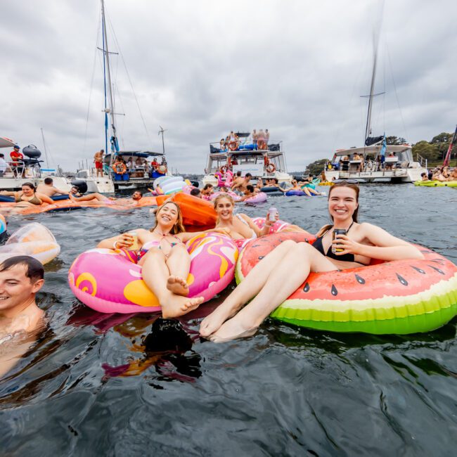 People enjoying a party in the water with boats anchored in the background. Some are floating on inflatable pool toys, including a large donut and a watermelon. The atmosphere is festive, with participants smiling and holding drinks, all part of The Yacht Social Club Event Boat Charters.