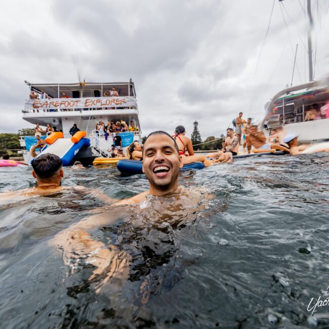 A man smiles while swimming in a body of water surrounded by others on pool floats and boats. Behind him, people are gathered on the deck of a large boat named "Barefoot Explorer." The atmosphere appears lively and festive under a cloudy sky, typical for Boat Parties Sydney The Yacht Social Club.