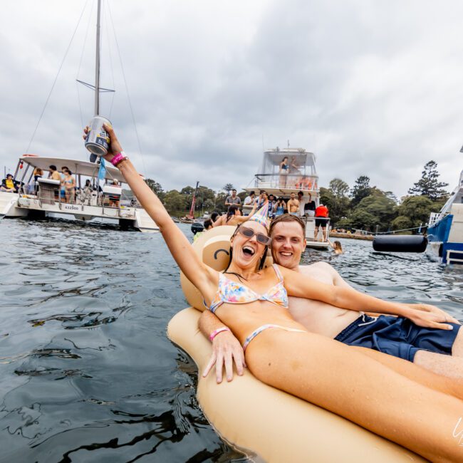 Two people relax on an inflatable lounge in the water, smiling and holding up drinks. Behind them are anchored boats with others enjoying a sunny day. Trees and a cloudy sky are visible in the background, creating a lively and festive atmosphere typical of The Yacht Social Club Sydney Boat Hire events.