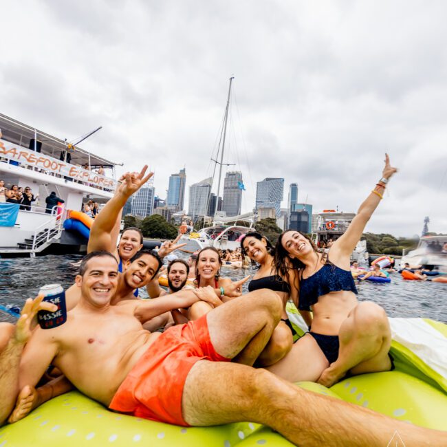 A group of cheerful people enjoying a day on the water, gathered on a large inflatable raft. They are posing for the camera against a backdrop of yachts and city skyline. Some are holding drinks, and everyone looks happy and relaxed under the cloudy sky with Sydney Harbour Boat Hire The Yacht Social Club.