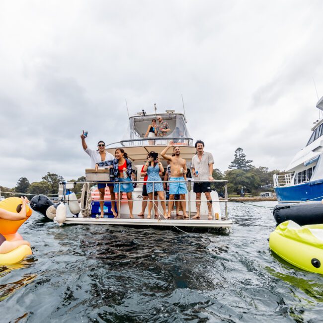 A lively scene on the water with a group of people enjoying a sunny day. Several individuals are on a luxury yacht from The Yacht Social Club, engaging and waving, while others float nearby on inflatable pool toys. Surrounding boats and a cloudy sky provide a picturesque backdrop.