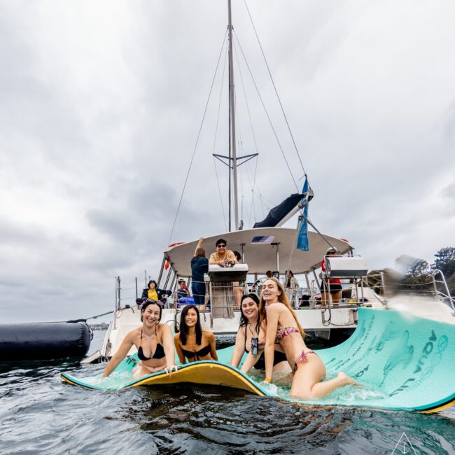 A group of four young women sits and lays on a floating mat in the water near a boat. They are smiling and wearing swimwear. The boat is anchored with a few people visible on it. The sky is overcast, and the water is calm. "Boat Parties Sydney The Yacht Social Club" is written in the bottom right corner.