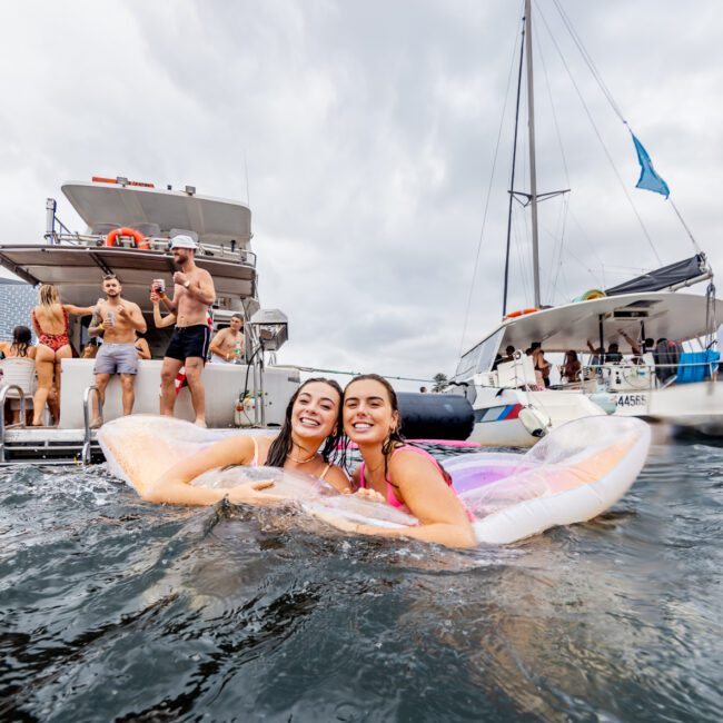 Two smiling women float on inflatable pool toys in the water, while a group of people socializes on a nearby boat. Another boat with a flag is anchored nearby. The sky is cloudy, and the scene looks like a lively event hosted by The Yacht Social Club, encapsulating the essence of Boat Parties Sydney.