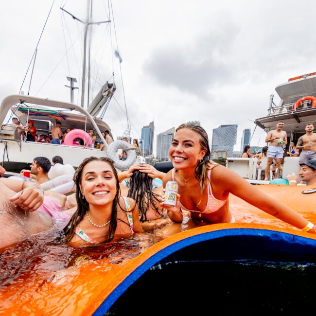 A lively group enjoys a party on a boat in a marina. Two women and a man, all in swimsuits, smile while lounging on an inflatable mat in the water. More people can be seen on boats in the background, with a cityscape visible under a cloudy sky. Experience it all with Sydney Harbour Boat Hire The Yacht Social Club.