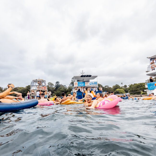 A group of people float on inflatable rafts and tubes in the water near two anchored boats. The boats, part of The Yacht Social Club Sydney Boat Hire, are decorated with banners and people are socializing. The sky is overcast, and in the background are trees and more boats.