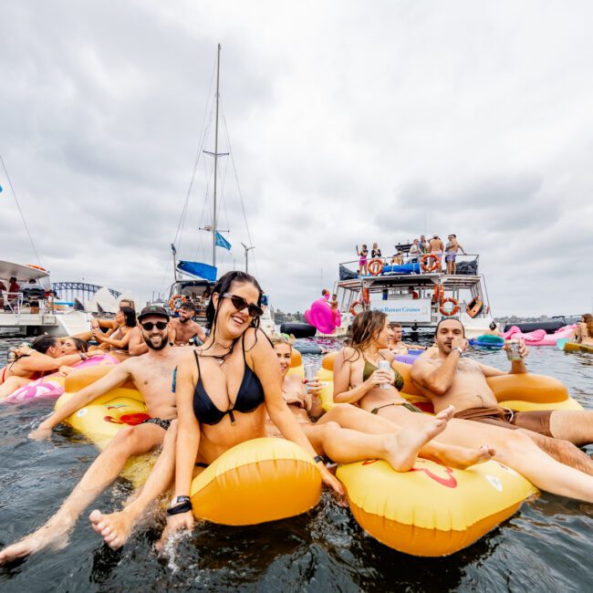 A group of people in swimwear enjoys a sunny day on the water, floating on yellow inflatable rafts. Several boats, part of The Yacht Social Club, are anchored in the background under a cloudy sky. Everyone appears relaxed and having fun.