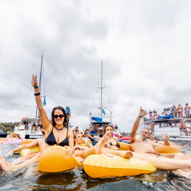 A group of people are floating on inflatable loungers in the water, enjoying a yacht party. One woman in a bikini raises her arm, smiling at the camera. Several boats and more people are visible in the background under a cloudy sky at The Yacht Social Club Event Boat Charters.