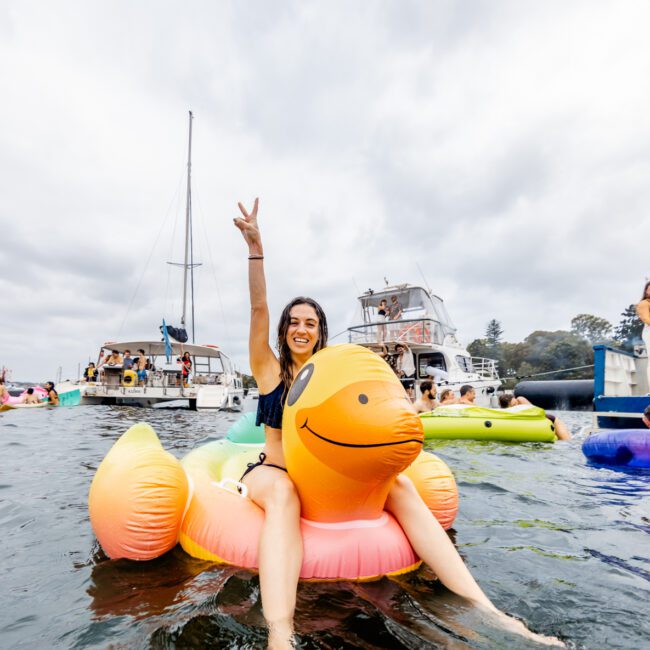 A person sits on an inflatable duck in the water, raising one arm in a peace sign. Several boats and people on inflatables are in the background. The sky is cloudy, and there are trees and buildings along the shoreline. The watermark says "Boat Parties Sydney The Yacht Social Club.