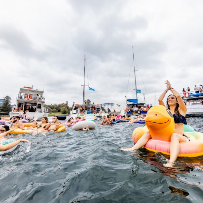 People enjoy a festive gathering on water, floating on various inflatable rafts and pool toys. A woman in sunglasses waves while sitting on an orange inflatable horse. Boats with more people and festive decorations are anchored nearby under a cloudy sky, courtesy of The Yacht Social Club Sydney Boat Hire.