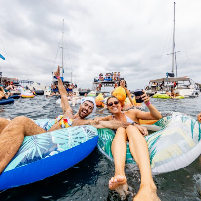 A group of people floating on inflatable tubes in the water, surrounded by boats. They appear to be celebrating, with one person raising a hand in the air and another holding a drink. The sky is overcast, and the scene is lively with numerous attendees having fun at Boat Parties Sydney The Yacht Social Club.