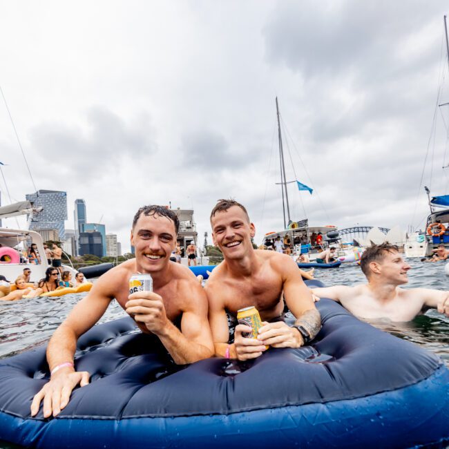 Two men smile and hold drinks while floating on an inflatable raft in a body of water with a group of people around them. Boats and the city skyline are in the background, under a cloudy sky. The atmosphere looks festive and lively, reminiscent of gatherings organized by Luxury Yacht Rentals Sydney.