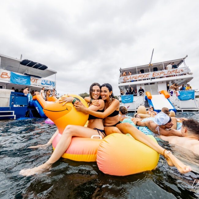 Two women are sitting on an inflatable float shaped like a cartoon character, smiling at the camera. They are surrounded by people in the water, with two large boats in the background. The scene appears to be a festive boat party hosted by The Yacht Social Club, adding to the vibrant atmosphere.