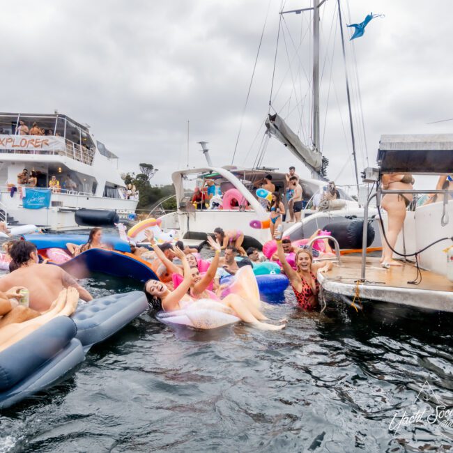 A lively scene of people enjoying a boat party hosted by The Yacht Social Club Sydney Boat Hire. The water is filled with colorful inflatable rafts and pool floats. Various individuals are swimming, laughing, and waving at the camera. Boats are anchored nearby, enhancing the festive and joyous atmosphere.