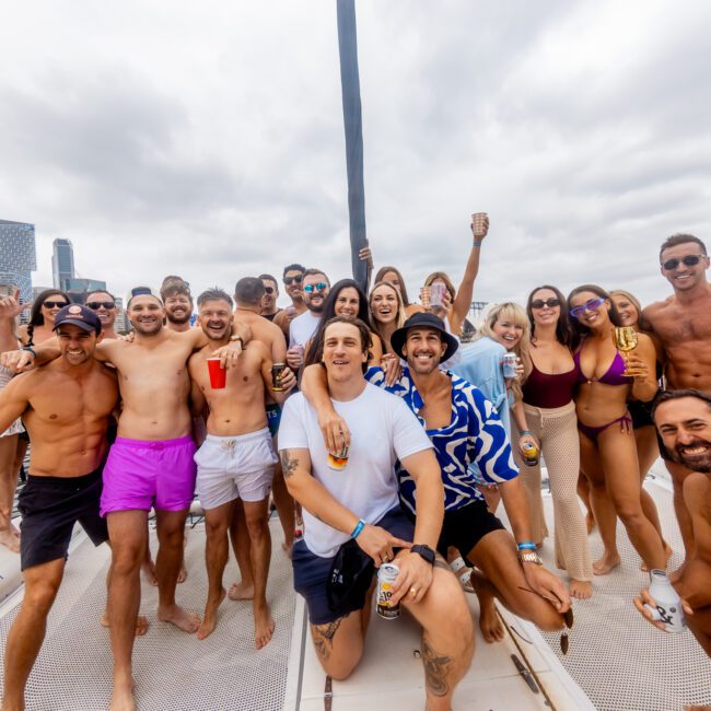 A cheerful group posing on a boat under cloudy skies, with men and women in swimwear, sunglasses, and summer attire. Some hold drinks, and everyone looks happy. Buildings and trees are visible in the background. The image has "Boat Parties Sydney The Yacht Social Club" logo in the bottom right corner.