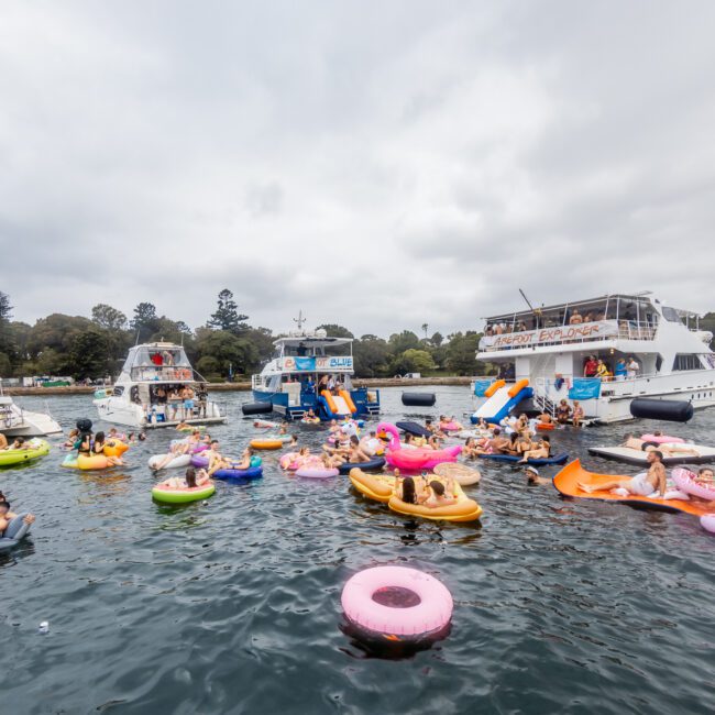 A lively scene of people on colorful inflatable rings floating on a body of water surrounded by various boats. The boats, hired from Sydney Harbour Boat Hire The Yacht Social Club, are anchored close together, creating a festive atmosphere. The sky is cloudy, and trees are visible in the background.